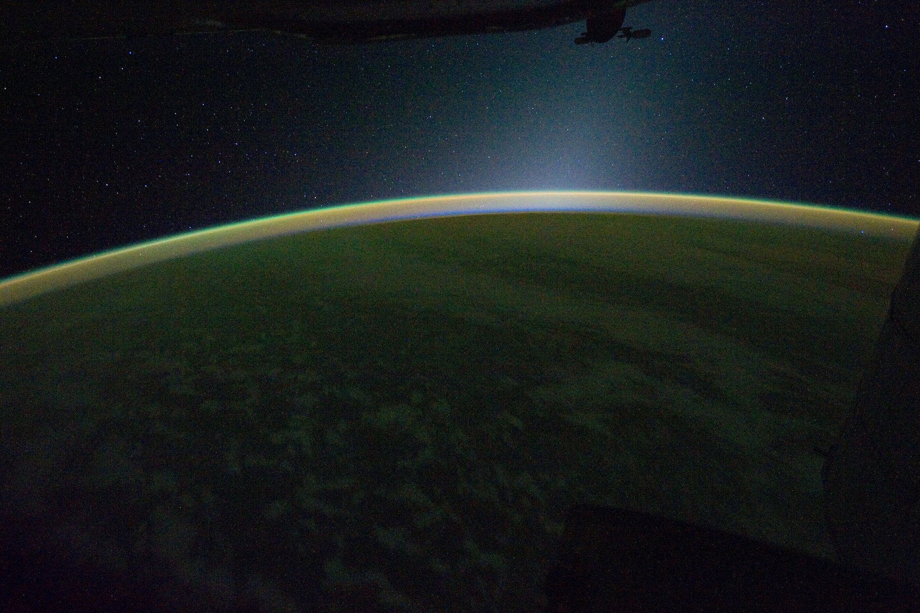 Thin glowing band of airglow along Earth's curved horizon at night, with stars above and dark clouds below