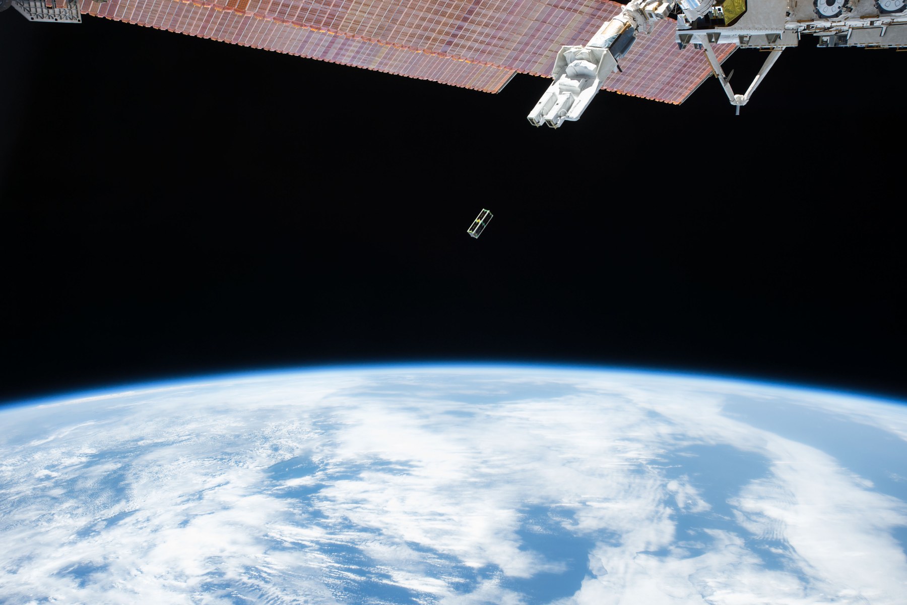 Small satellite released from the International Space Station drifting above a cloud-covered Earth
